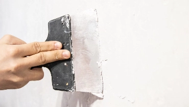 A close-up of a handyman's hand as they use a putty spatula to complete drywall repair for a white wall.