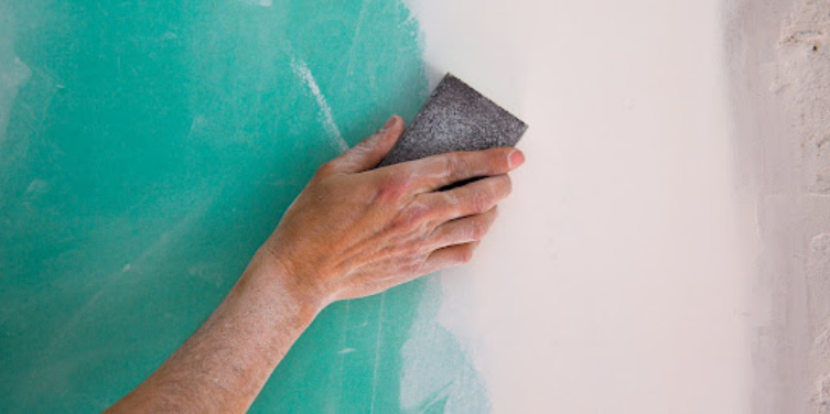 A hand holding a sanding brush as someone uses the brush to sand down the unpainted area of a wall where drywall repairs have been completed.