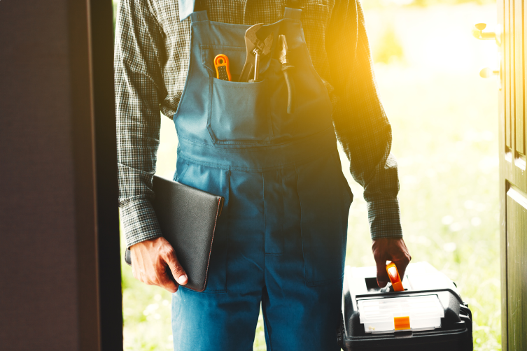 Handyman wearing overalls and standing in a doorway holding a toolbox, shown from mid-chest to just above the knee.