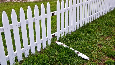 A white picket fence with a single picket broken off and lying on the ground before it’s been re-attached with fence repair service.