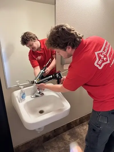 A Mr. Handyman service professional caulks the space where a white porcelain sink meets the wall.