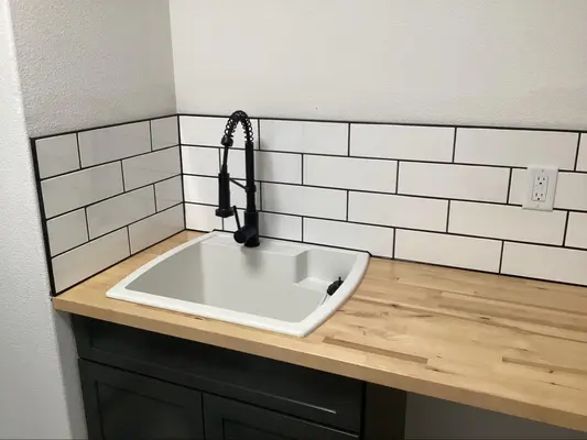 A white sink in a wooden countertop is surrounded by a white tiled backsplash that has been set with black grout.