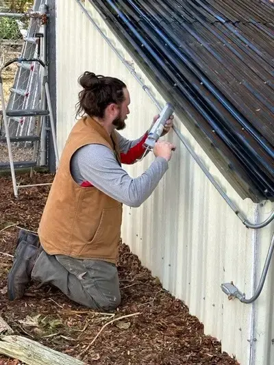 A Mr. Handyman service professional kneels down outside to put caulking between the siding and roof of a metal shed.