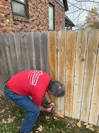 A Mr. Handyman service professional bends down to drill into a fence with an electric drill.