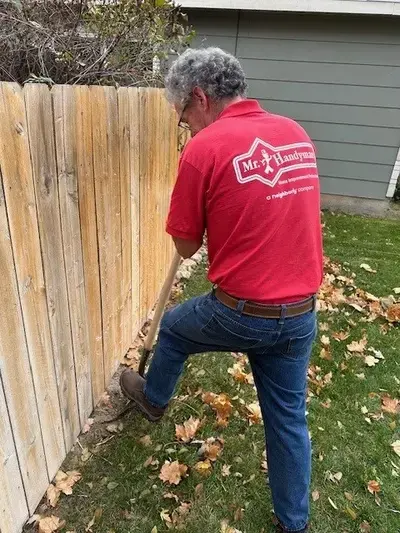 A Mr. Handyman service professional stands outside digging into the ground with a shovel at the base of a wooden fence.