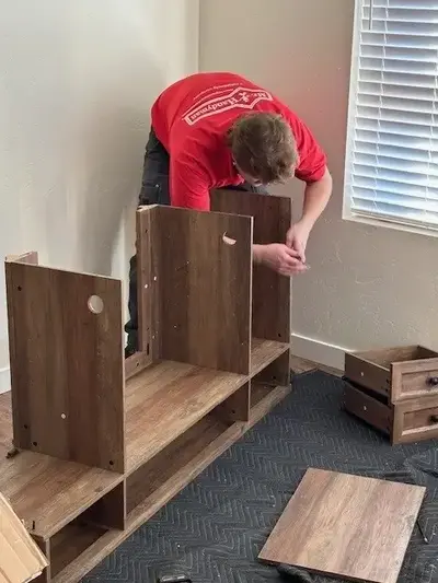 A Mr. Handyman service professional in a red Mr. Handyman shirt is assembling a large wooden desk.