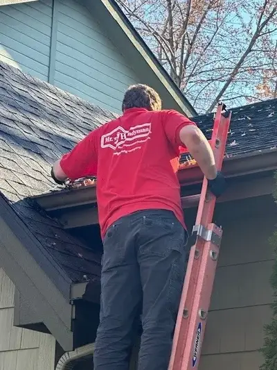 A Mr. Handyman service professional is standing on a red ladder cleaning out gutters on a black shingled roof.