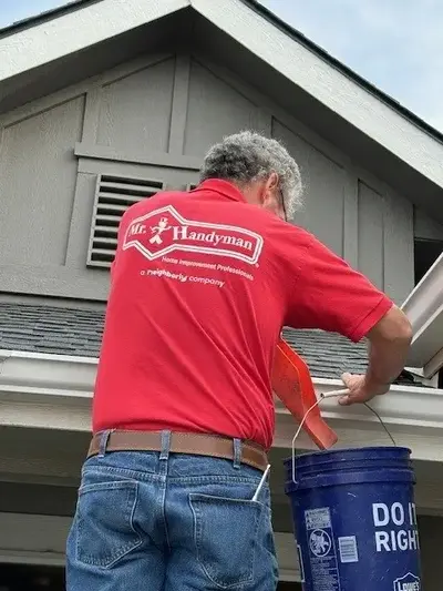 A Mr. Handyman service professional in a red Mr. Handyman shirt empties debris from a gutter on the roof into a blue bucket.