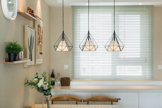 Three pendant lights hang over a dining room table. The room is tastefully decorated and there is a large window covered in blinds in the background.