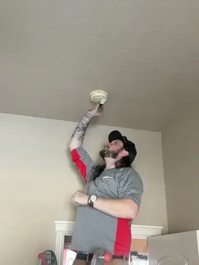 A Mr. Handyman service professional reaches up to change the batteries on a smoke detector.
