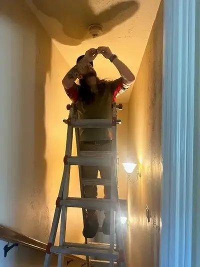 A Mr. Handyman service professional stands on a metal ladder propped up on stairs to change a hallway smoke detector.