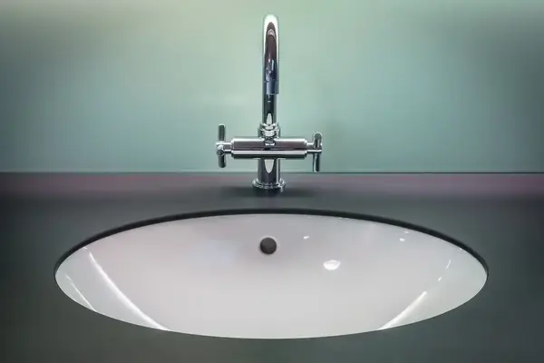 A closeup image of a white ceramic sink with a stainless steel faucet set into a dark countertop.