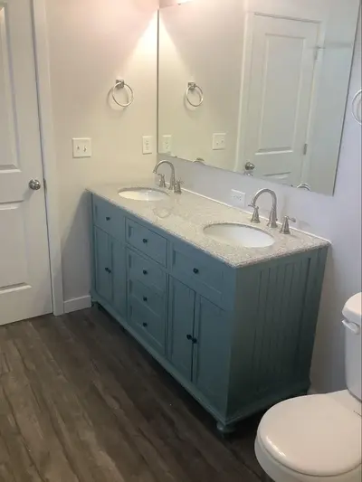 A dusty blue vanity with two sinks on top with stainless steel faucets and handles next to a toilet with a mirror above.