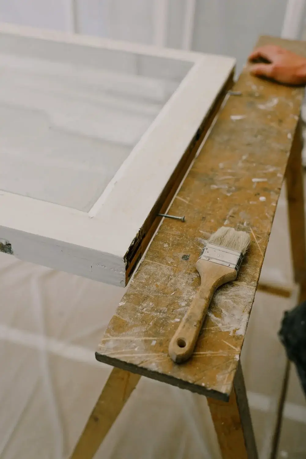 A window and a paintbrush are on a work bench. There is a hand on the workbench in the background.