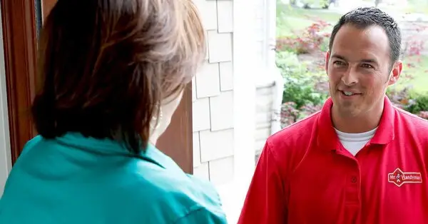 A smiling handyman wearing a Mr. Handyman uniform speaking with a homeowner at their front door.