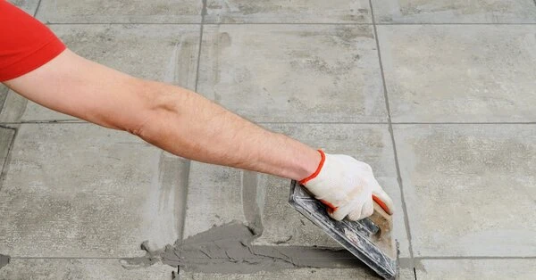 A handyman using a trowel to spread fresh grout into the gap between two floor tiles during an appointment for grout repair in Wichita, KS.