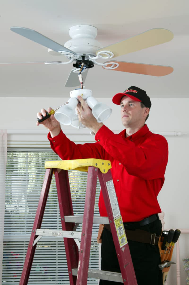 A handyman from Mr. Handyman standing on a ladder and installing a new ceiling fan in the living room of a home.