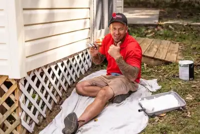 Mr. Handyman technician giving thumbs up sign while sitting on ground next to exterior of home working on painting project.