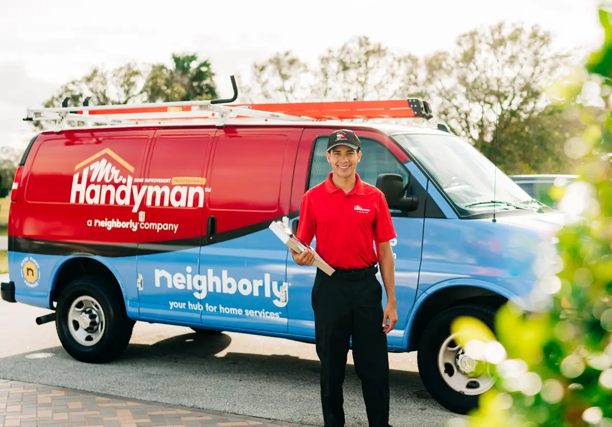 A Mr. Handyman technician standing in front of his company van holding a clipboard.