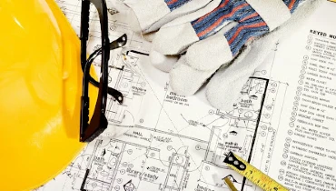 A hardhat, safety glasses, work gloves and the end of a tape measurer lying on top of detailed home remodeling plans.