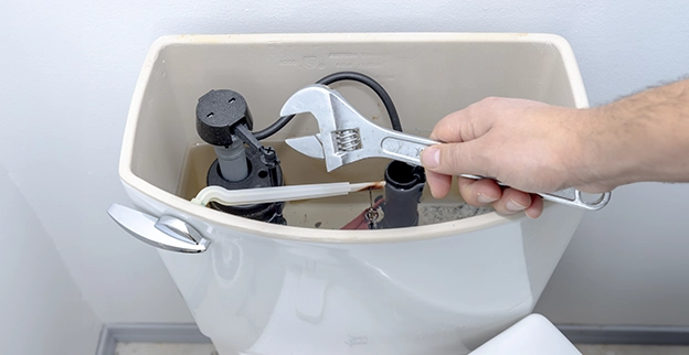 A hand using an adjustable wrench to repair the internal components of a toilet tank.