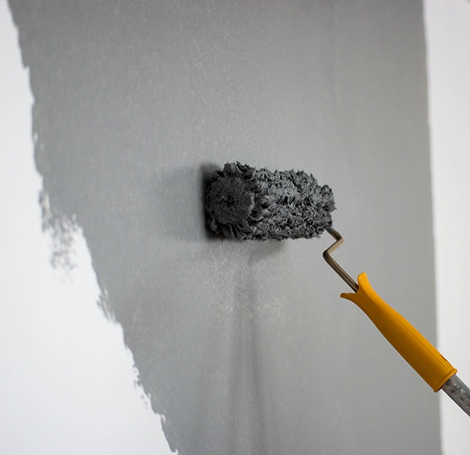 A paint roller with a yellow handle applies grey paint to a wall.