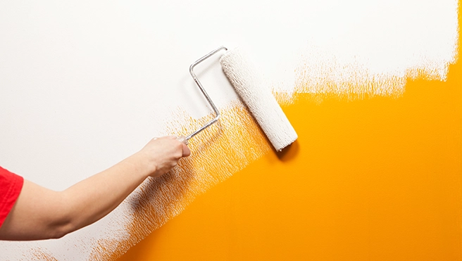 A hand painting a wall with a roller, applying vibrant orange paint.