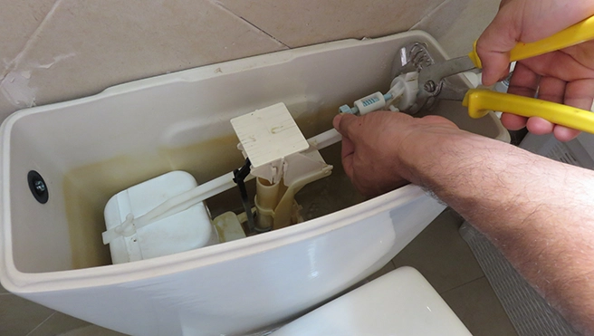A person's hand uses pliers to repair the mechanism inside a toilet tank.