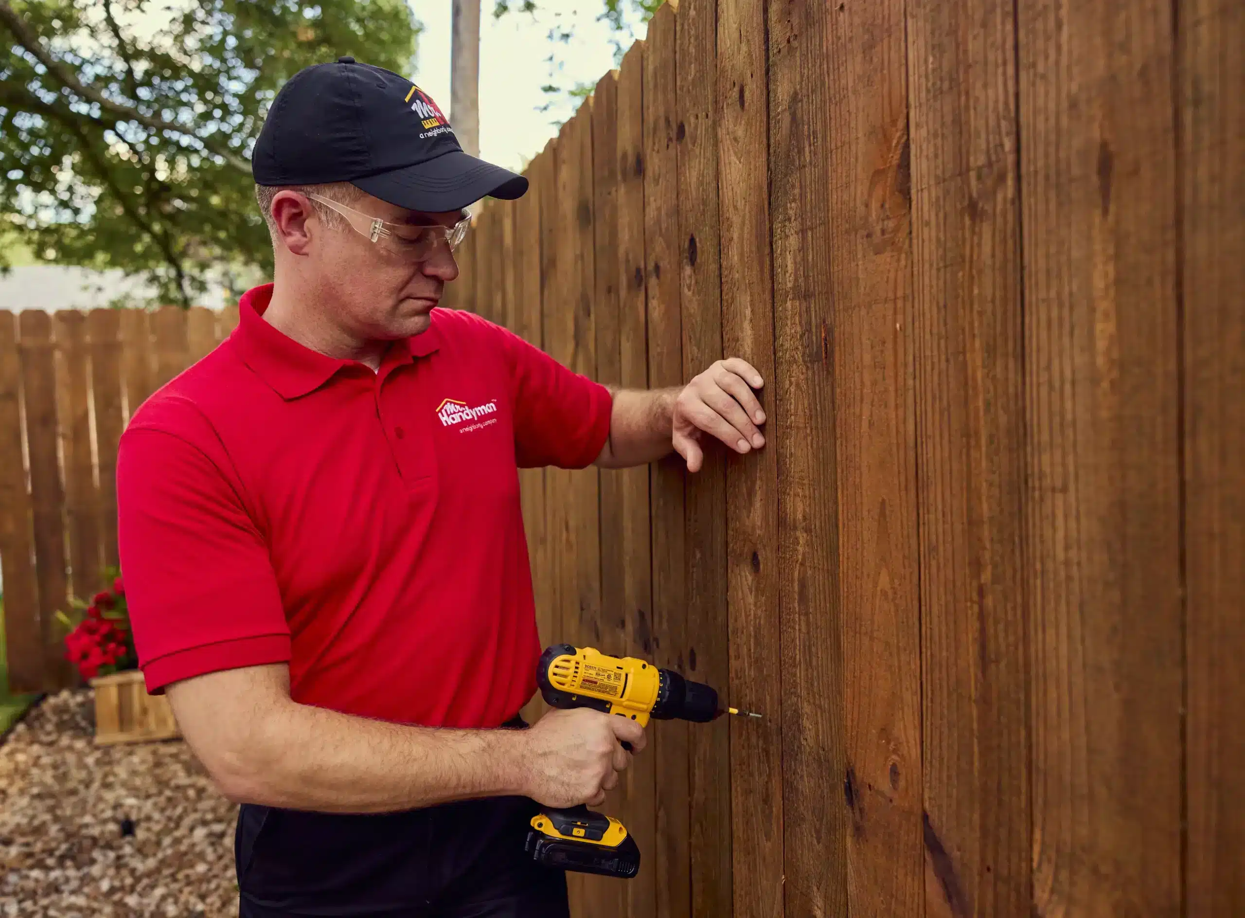 A uniformed Mr. Handyman service professional using a power drill to repair a wooden outdoor fence.