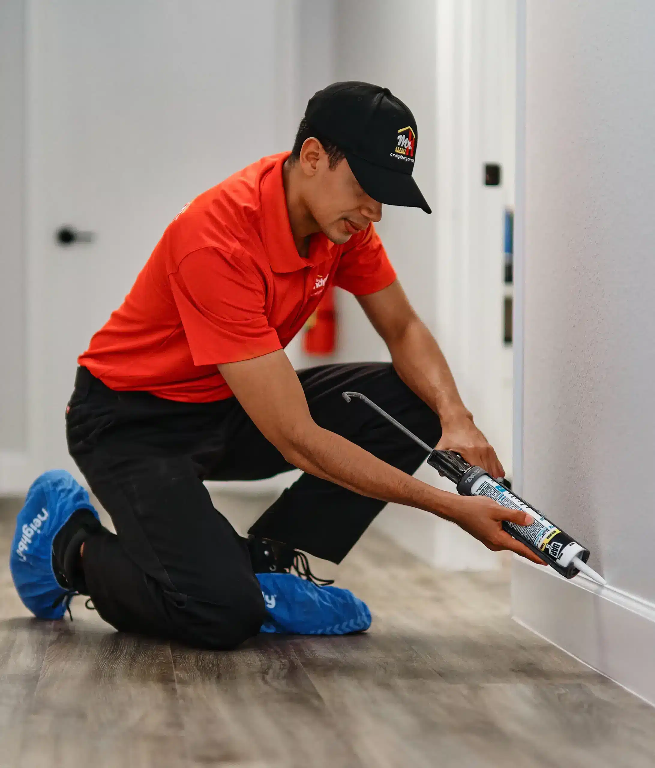 A uniformed Mr. Handyman service professional applying caulk to interior baseboard trim.