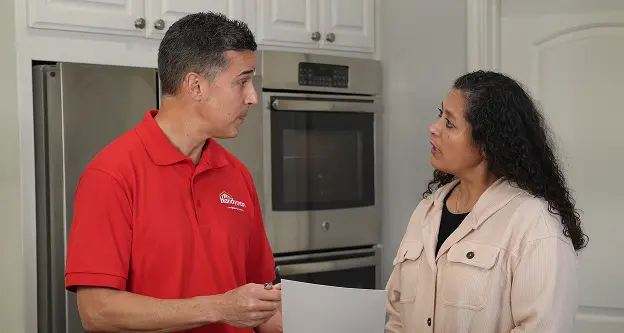 A uniformed Mr. Handyman service professional discussing a project estimate with a homeowner in her kitchen.
