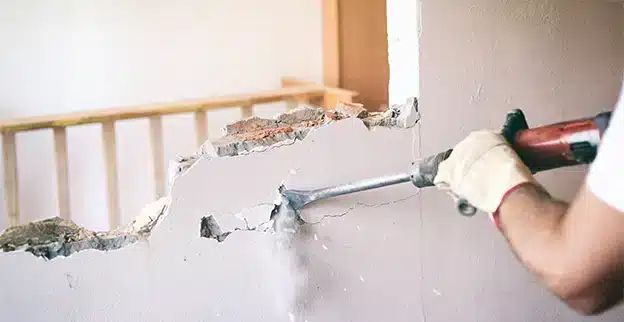 Mr. Handyman professional wearing gloves uses a jackhammer to break through a wall, creating dust and debris.