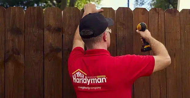 A uniformed My Handyman service professional using a power drill to repair a wooden outdoor fence.