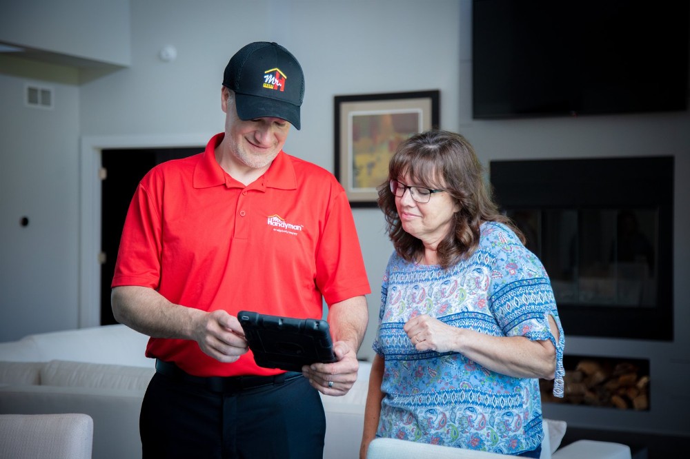 Mr. Handyman technician in a red shirt showing project details on a tablet to a smiling homeowner in her living room