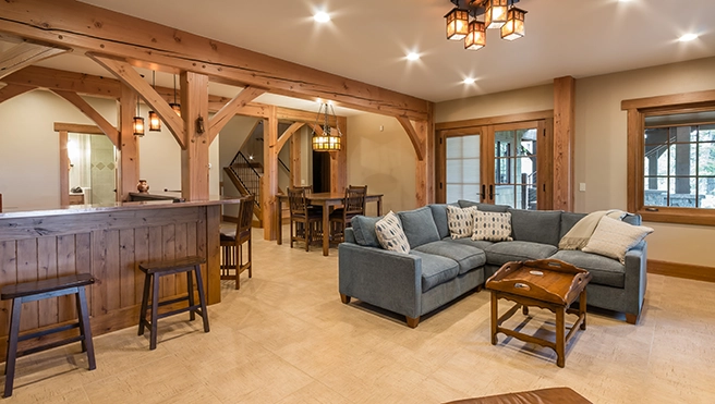 Cozy living space with a blue sectional sofa, wooden bar, dining area, and large windows, featuring rustic beams and warm lighting.