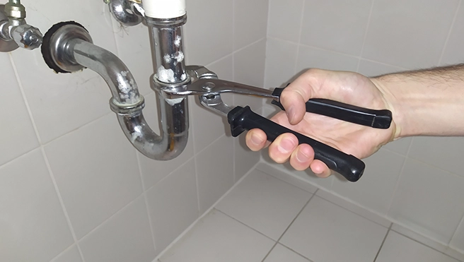 A hand holding pliers adjusts a plumbing pipe under a sink, showcasing DIY plumbing repair work in a bathroom setting.