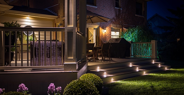 A cozy evening scene of a well-lit porch with furniture, a grill, and blooming flowers.