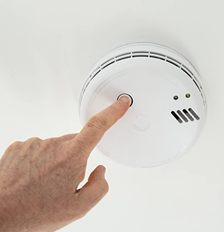 A hand pressing the test button on a white smoke detector mounted on a ceiling.