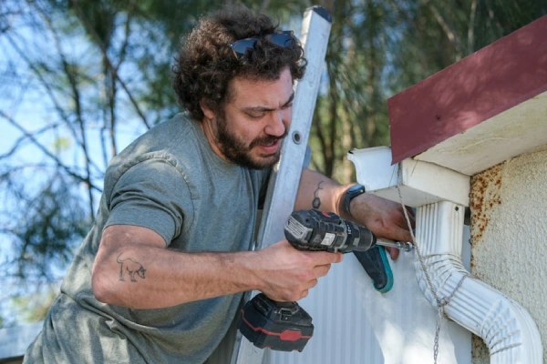 Someone stands on a ladder and uses a drill to fix a gutter on their Idaho home.
