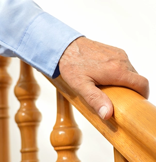 A close-up of an elderly hand resting on a polished wooden handrail, wearing a light blue shirt.