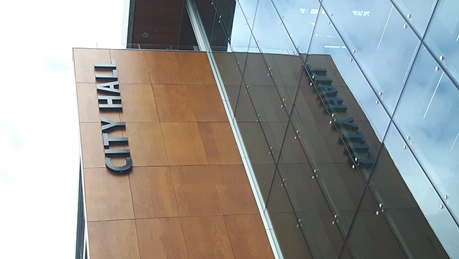 A modern City Hall building with wooden panels and glass façade, featuring the bold text.