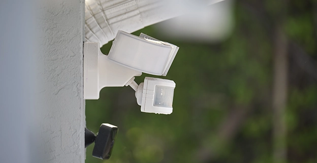 A close-up of a white motion sensor light fixture installed on a textured wall surrounded by greenery.