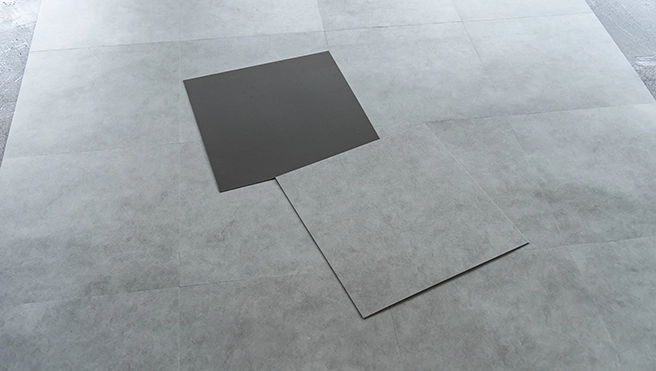 A light gray textured floor tiles with two dark gray square samples placed on it for comparison.