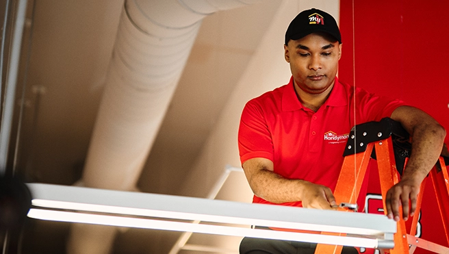 A handyman installing a light fixture while standing on a ladder in a bright, spacious indoor setting.