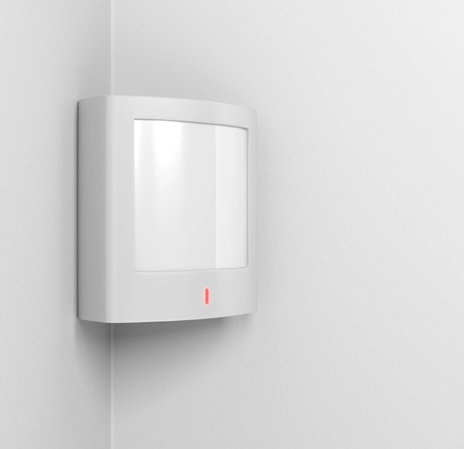 A white motion sensor installed on a corner wall featuring a small red indicator light on a smooth wall surface.