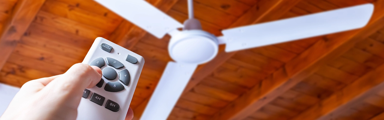 A remote control aimed at a ceiling fan with white blades, set against a wooden ceiling background.