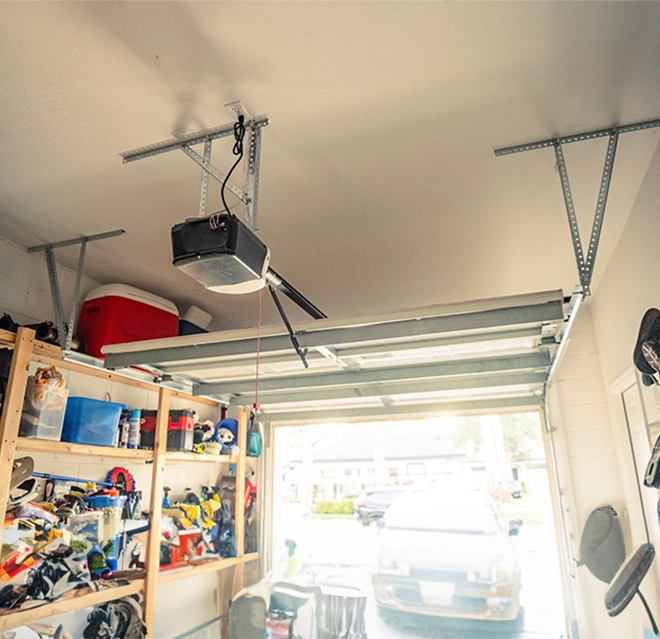 A garage interior a mounted garage door opener and shelves filled with tools, toys, and a visible car outside.