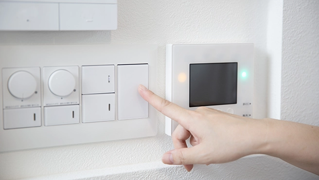 A hand pressing a white light switch next to a digital thermostat.