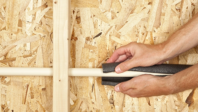 A person's hands wrap insulation around a white pipe on an oriented strand board wall.