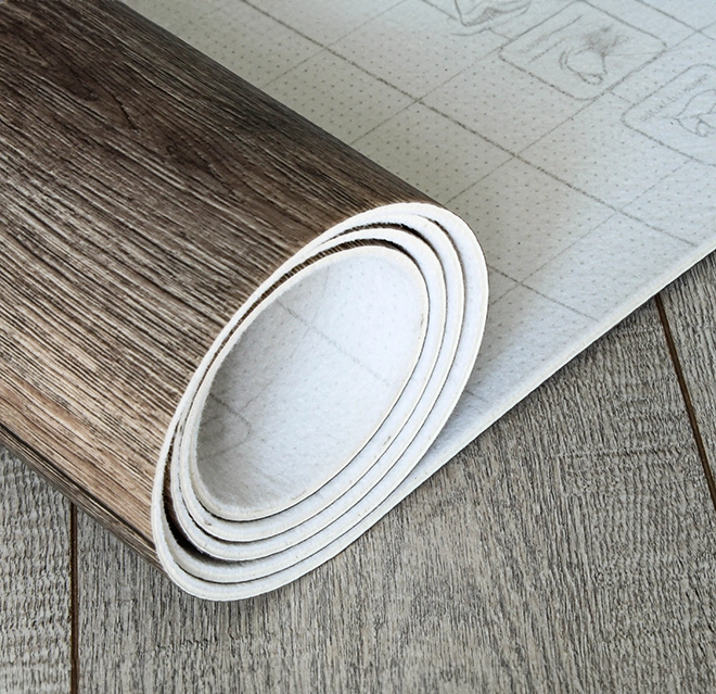 A rolled-up vinyl floor covering with a wood-like texture, partially unrolled onto a wooden surface.